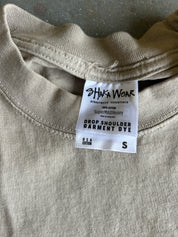 Close-up of a light beige crewneck garment dye tag from Shaka Wear, showing the brand name and size information on the inside collar of a cotton shirt.