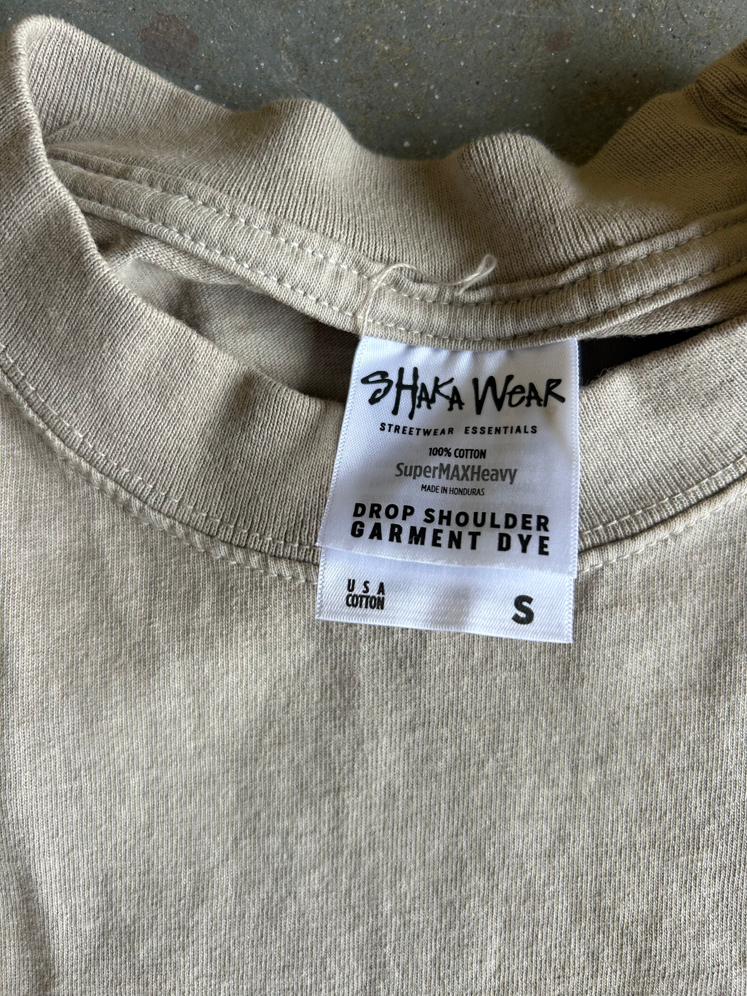 Close-up of a light beige crewneck garment dye tag from Shaka Wear, showing the brand name and size information on the inside collar of a cotton shirt.