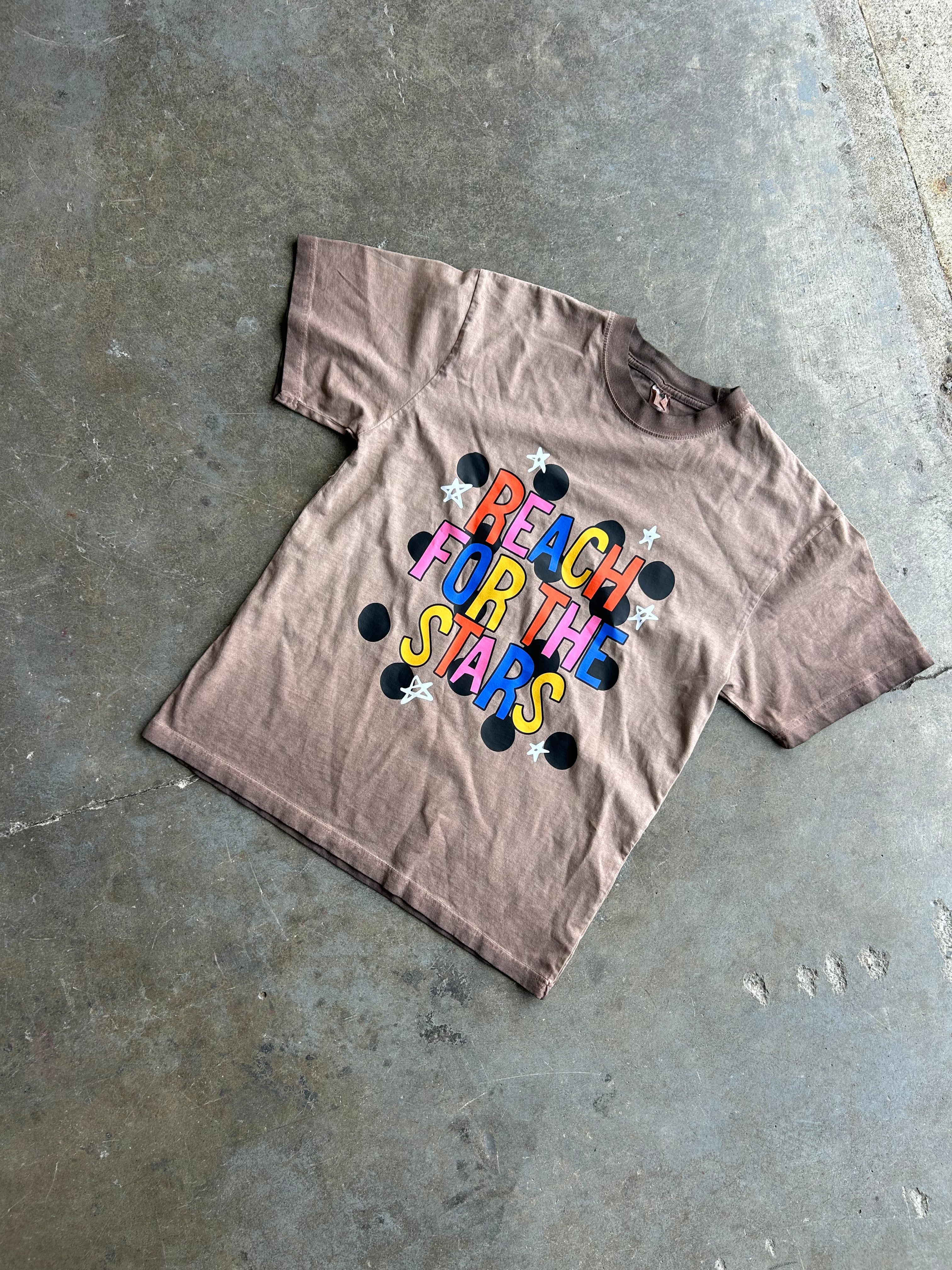 A faded brown short-sleeve t-shirt with a colorful retro graphic reading 'REACH FOR THE STARS' in rainbow letters with black and white stars, laid flat on a concrete floor.