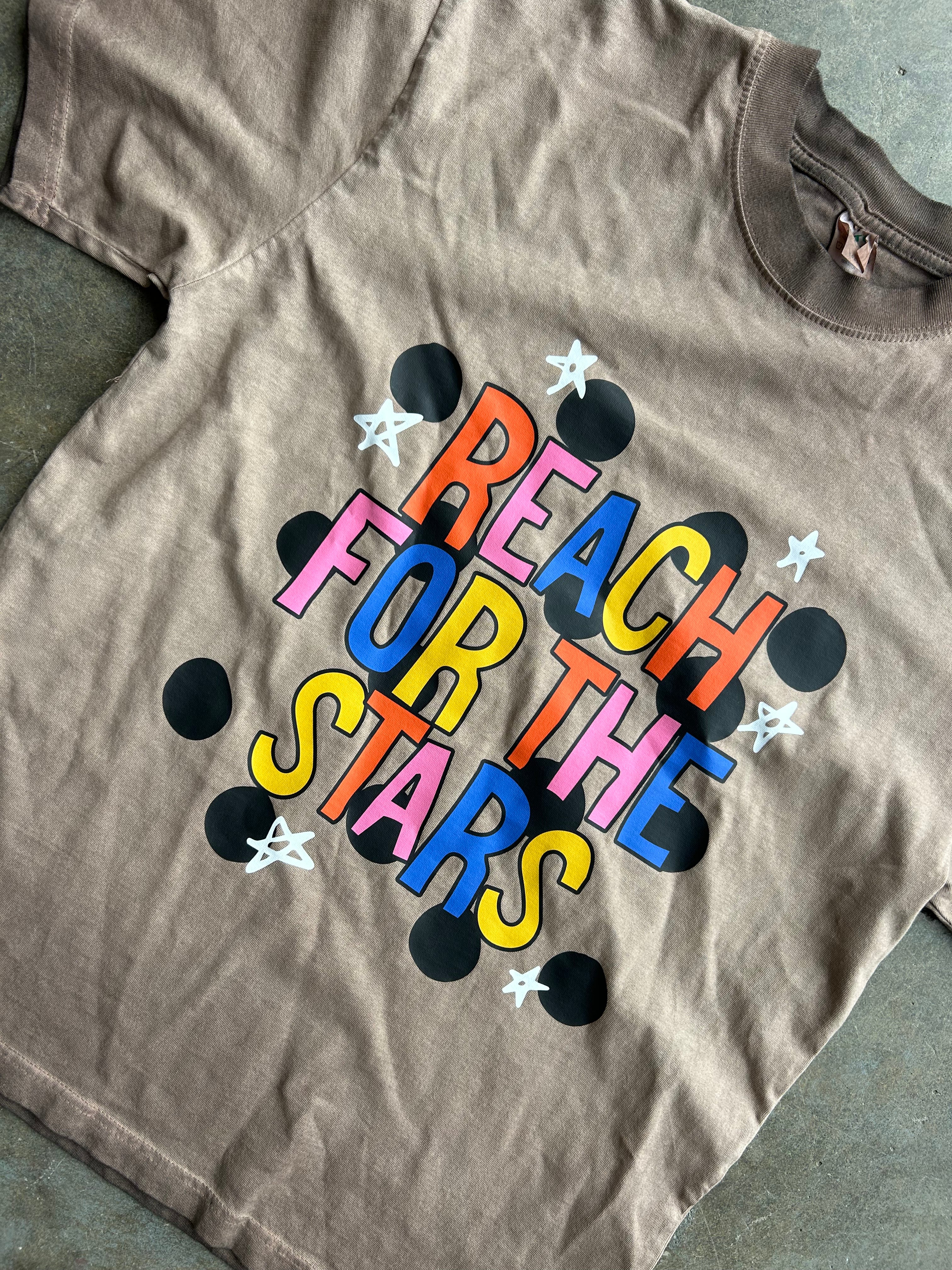 A taupe brown short-sleeve t-shirt laid flat on a gray concrete surface, featuring a colorful graphic that reads 'REACH FOR THE STARS' in multicolored letters, surrounded by black dots and white stars.