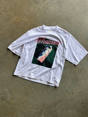 A white short-sleeve graphic t-shirt laid flat on a textured concrete floor. The shirt features a large front graphic with the word 'Whatever!' in pink script above a square image of a person in a pink striped swimsuit in a green forest.