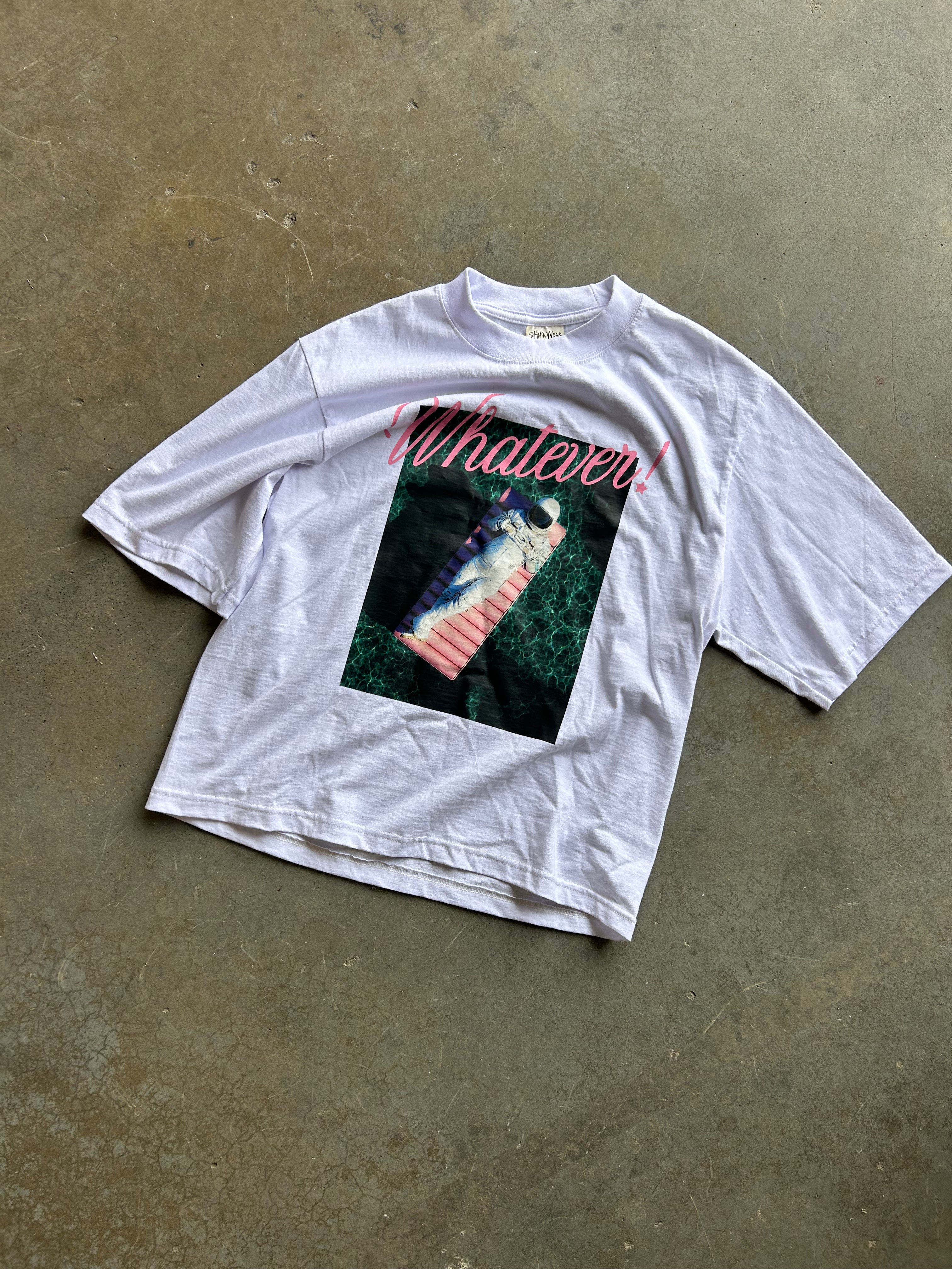 A white short-sleeve graphic t-shirt laid flat on a textured concrete floor. The shirt features a large front graphic with the word 'Whatever!' in pink script above a square image of a person in a pink striped swimsuit in a green forest.