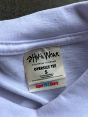 Close-up of the inside collar of a light lavender oversized t-shirt, showing a cream-colored brand label for 'Shaka Wear' with 'Streetwear Essentials' and 'Oversize Tee' printed on it.