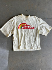 A cream-colored short-sleeve t-shirt laid flat on a gray concrete floor, featuring a red and green graphic on the chest that reads 'RUNNING LATE AGAIN'.