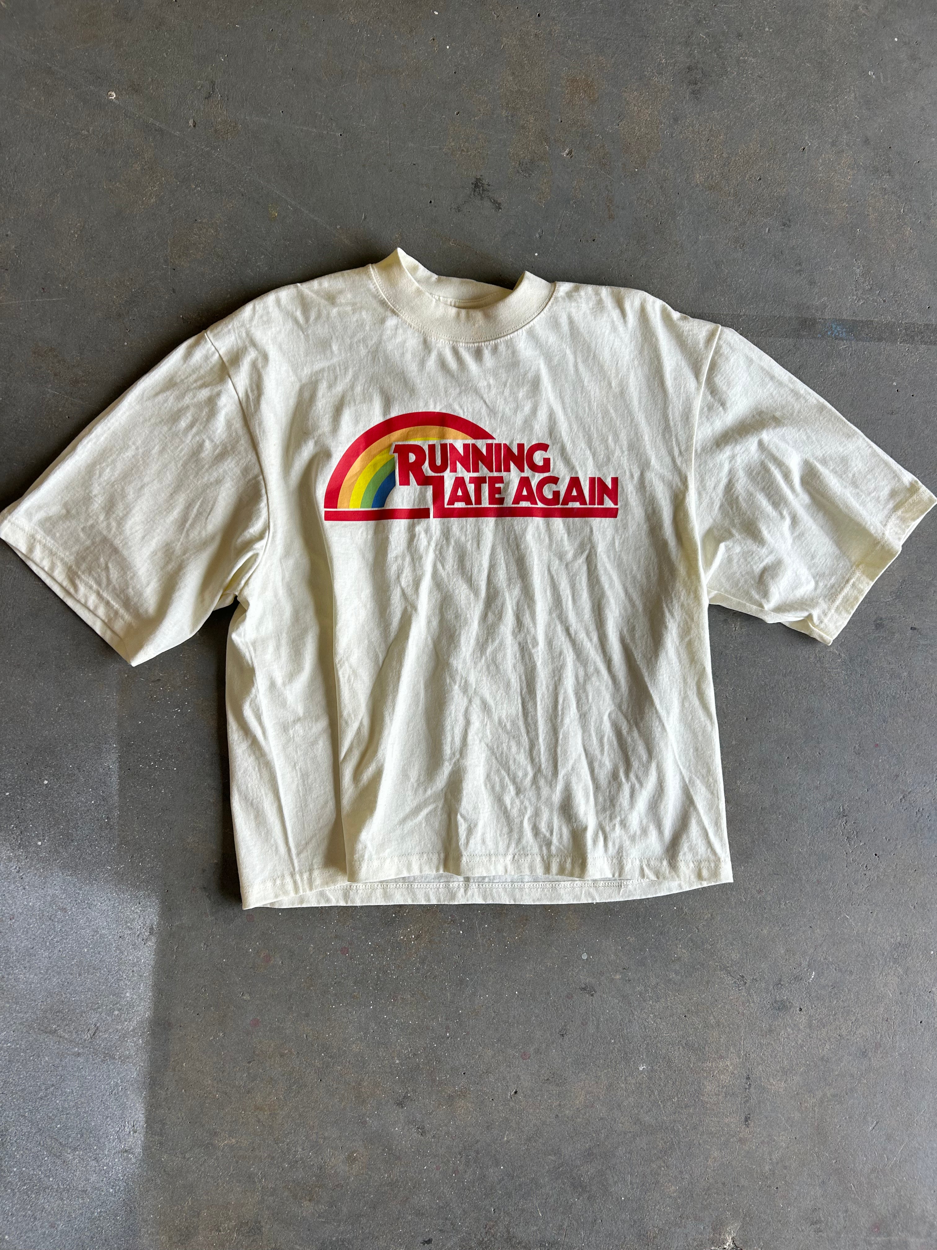 A cream-colored short-sleeve t-shirt laid flat on a gray concrete floor, featuring a red and green graphic on the chest that reads 'RUNNING LATE AGAIN'.