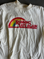 Close up of a cream-colored short-sleeve t-shirt laid flat on a gray concrete floor, featuring a red and green graphic on the chest that reads 'RUNNING LATE AGAIN'.