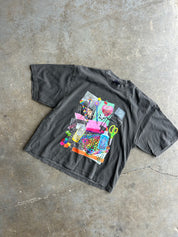 A faded black graphic t-shirt laid flat on a textured concrete floor. The shirt features a large, colorful collage print on the front depicting various craft supplies like scissors, beads, and paintbrushes.