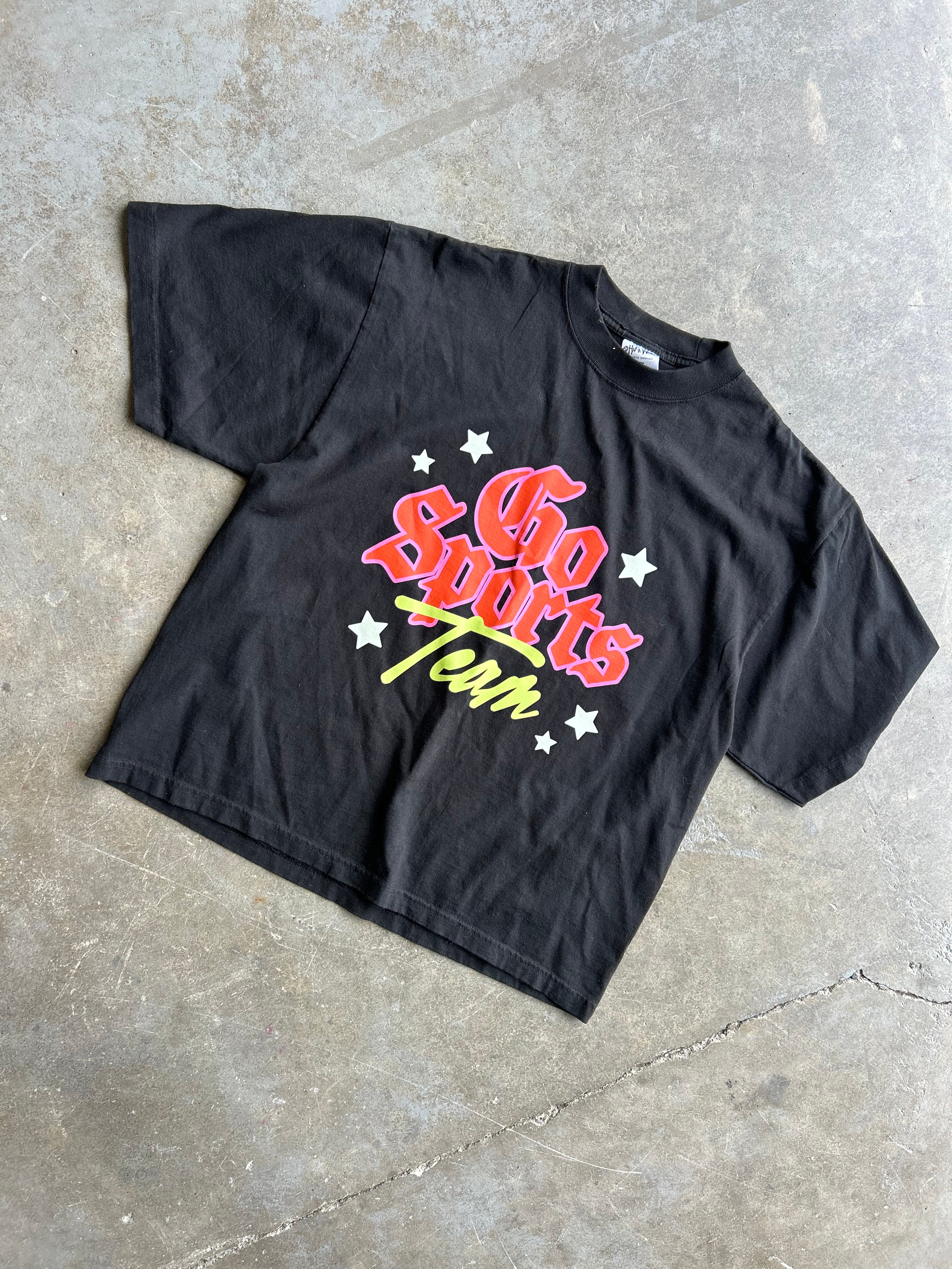 A black short-sleeve graphic t-shirt laid flat on a textured concrete floor. The shirt features a retro-style 'Go Sports Team' graphic in red, yellow, and white with star accents.