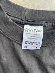 Close-up of a dark gray garment tag reading 'SHAKA WEAR' and 'Drop Shoulder Garment Dye' on a dark charcoal sweatshirt, with a concrete surface visible in the background.