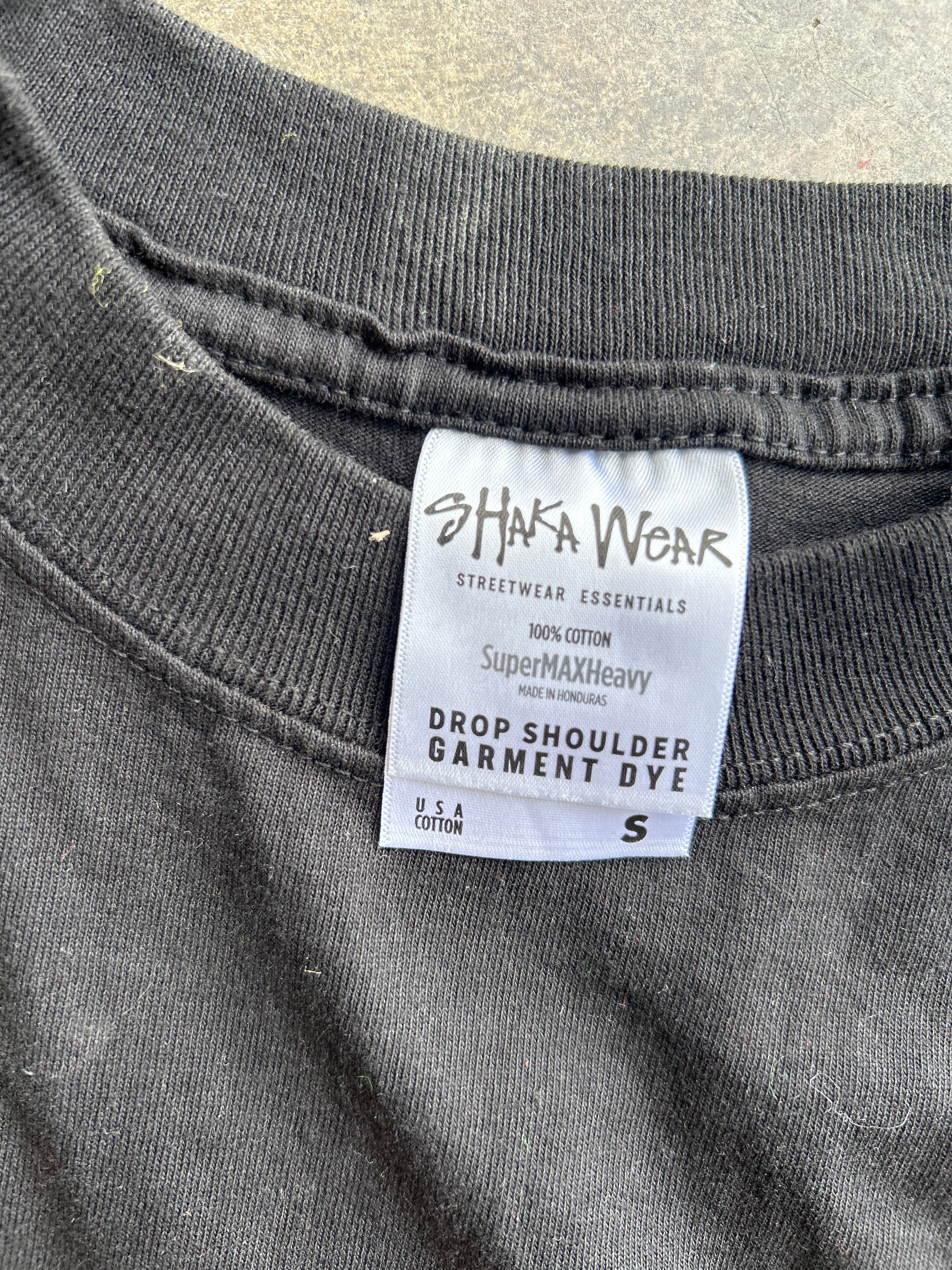 Close-up of a dark gray garment tag reading 'SHAKA WEAR' and 'Drop Shoulder Garment Dye' on a dark charcoal sweatshirt, with a concrete surface visible in the background.