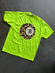 A bright neon yellow-green t-shirt laid flat on a dark gray concrete floor. The shirt features a circular graphic on the chest with a leopard print border and a white billiard 8-ball in the center.