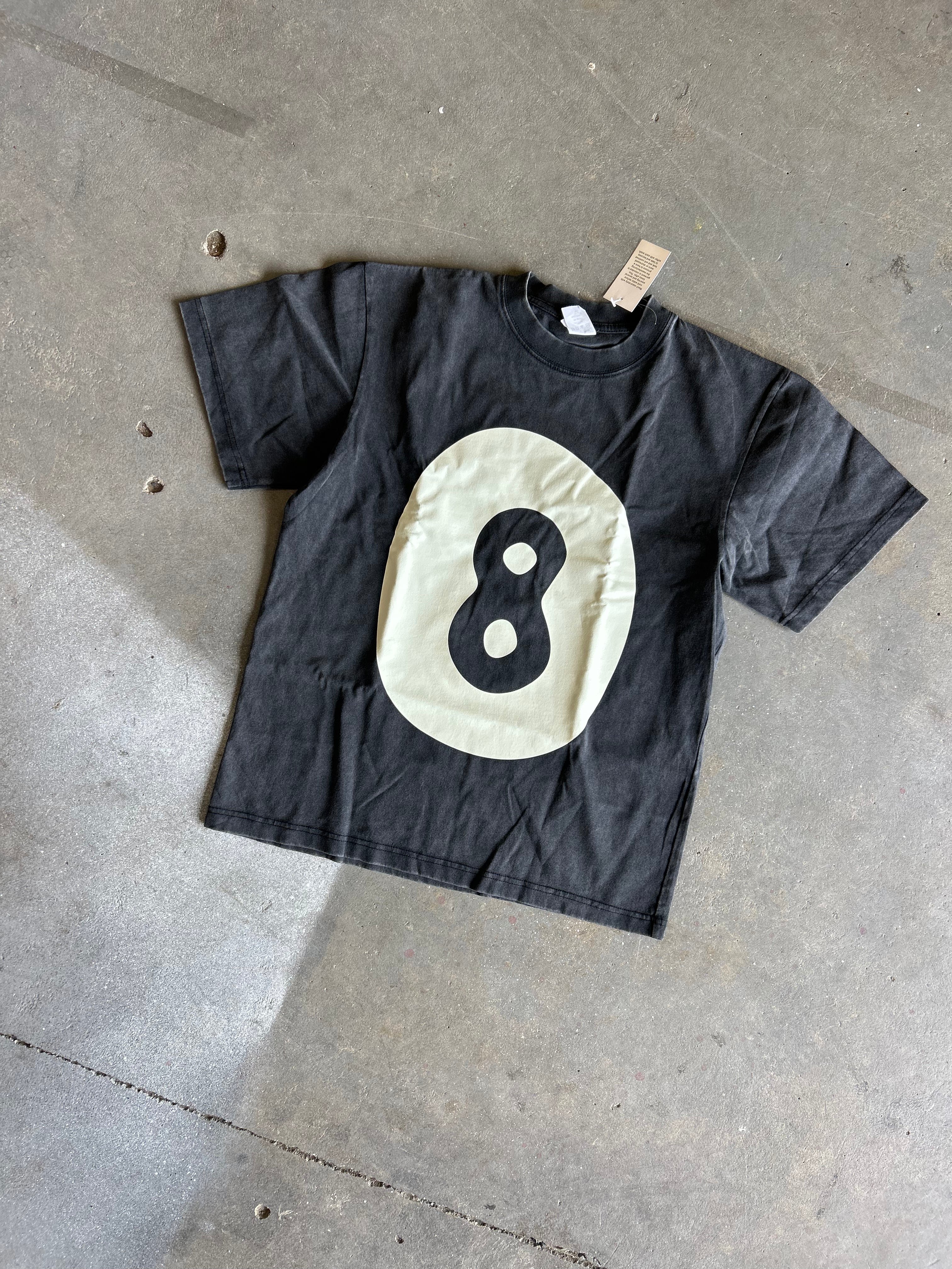 A black short-sleeve t-shirt with a large cream-colored oval graphic on the front featuring a black number 8, laid flat on a gray concrete floor.