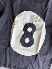 A close-up of a dark charcoal gray t-shirt featuring a large cream-colored circular graphic with a black stylized number 8 printed inside.