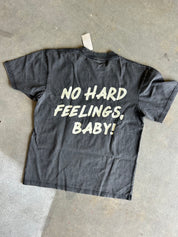 A faded black graphic t-shirt laid flat on a concrete floor, featuring the text 'NO HARD FEELINGS, BABY!' printed in a distressed cream-colored font on the back.