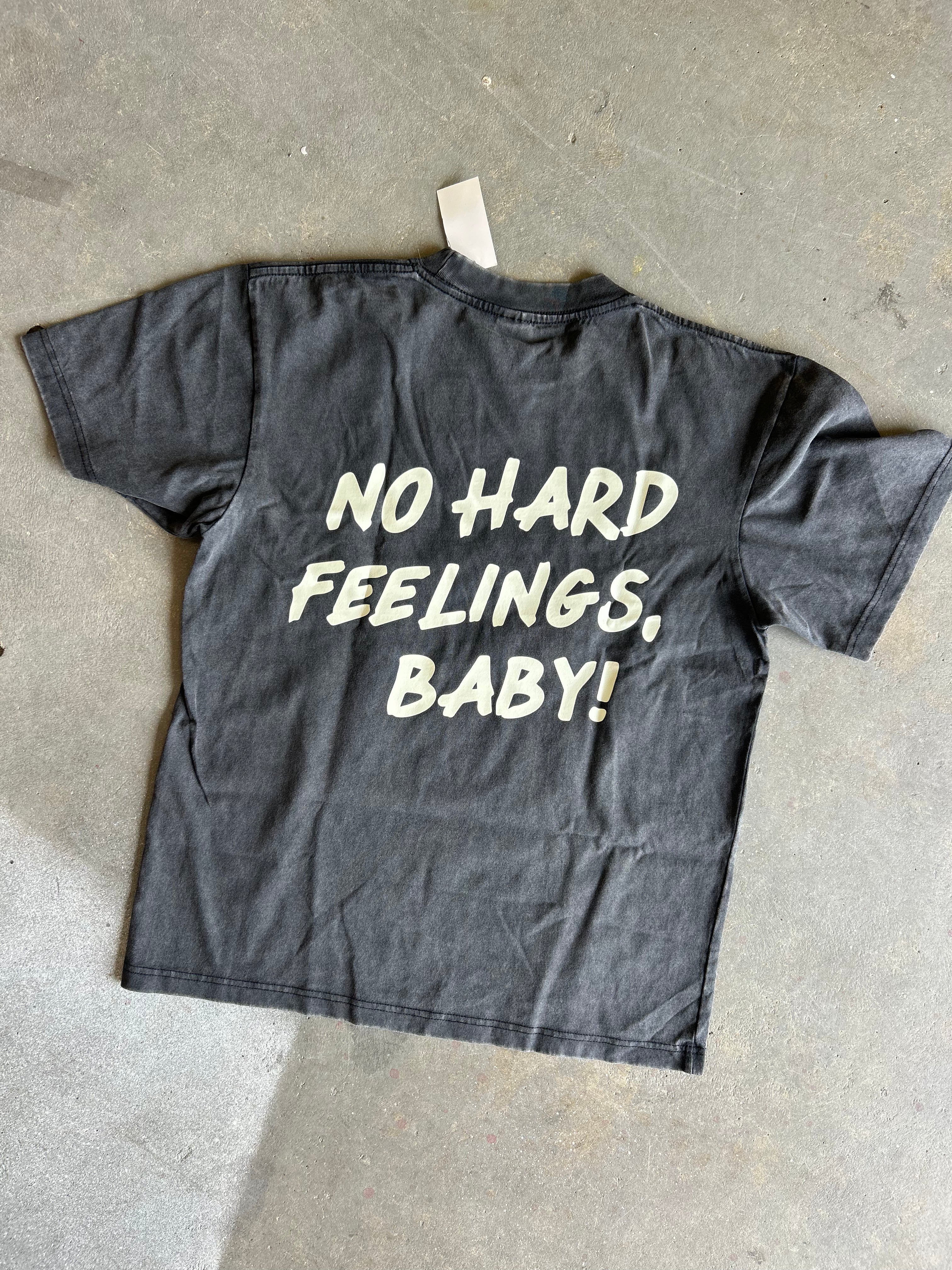 A faded black graphic t-shirt laid flat on a concrete floor, featuring the text 'NO HARD FEELINGS, BABY!' printed in a distressed cream-colored font on the back.