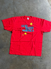 A red short-sleeve t-shirt laid flat on a gray concrete floor, featuring the text 'GOD BLESS AMERICA' in blue and yellow stars.