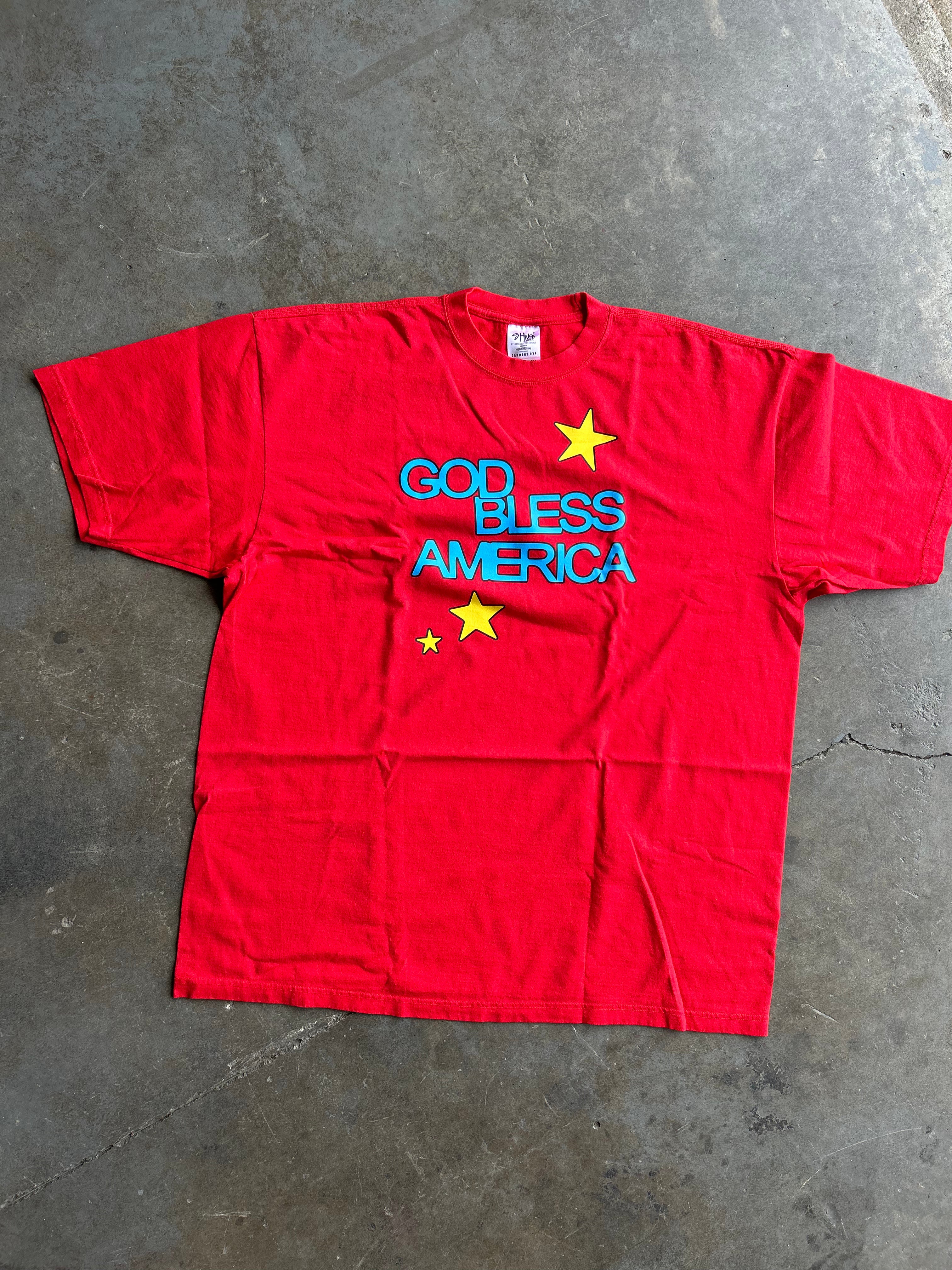 A red short-sleeve t-shirt laid flat on a gray concrete floor, featuring the text 'GOD BLESS AMERICA' in blue and yellow stars.