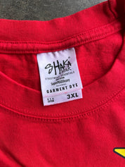 Close-up of a bright red t-shirt showing the inner neck label which reads 'SHAKA STREETWEAR ESSENTIALS' and 'GARMENT DYE 3XL'.