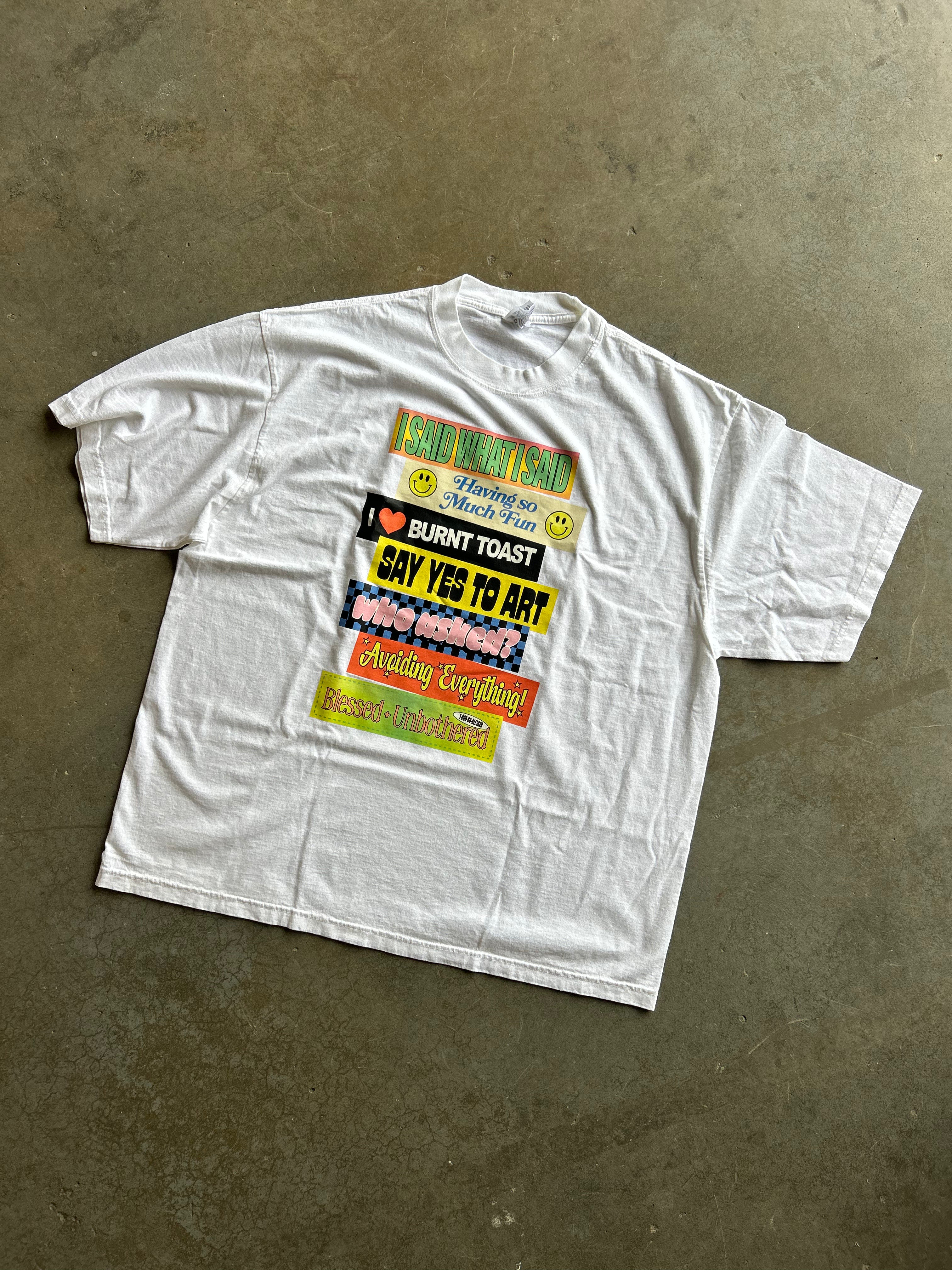 A white short-sleeve graphic t-shirt laid flat on a textured concrete floor. The front of the shirt features a colorful collage of text stickers in red, yellow, green, and black.