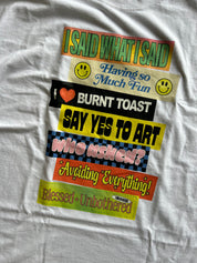 A close-up of a white t-shirt featuring a stacked graphic print with colorful retro-style stickers and bold text including 'I SAID WHAT I SAID', 'Having so Much Fun', 'I ❤— BURNT TOAST', 'SAY YES TO ART', 'who asked?', 'Avoiding Everything!', and 'Blessed Unbothered'.