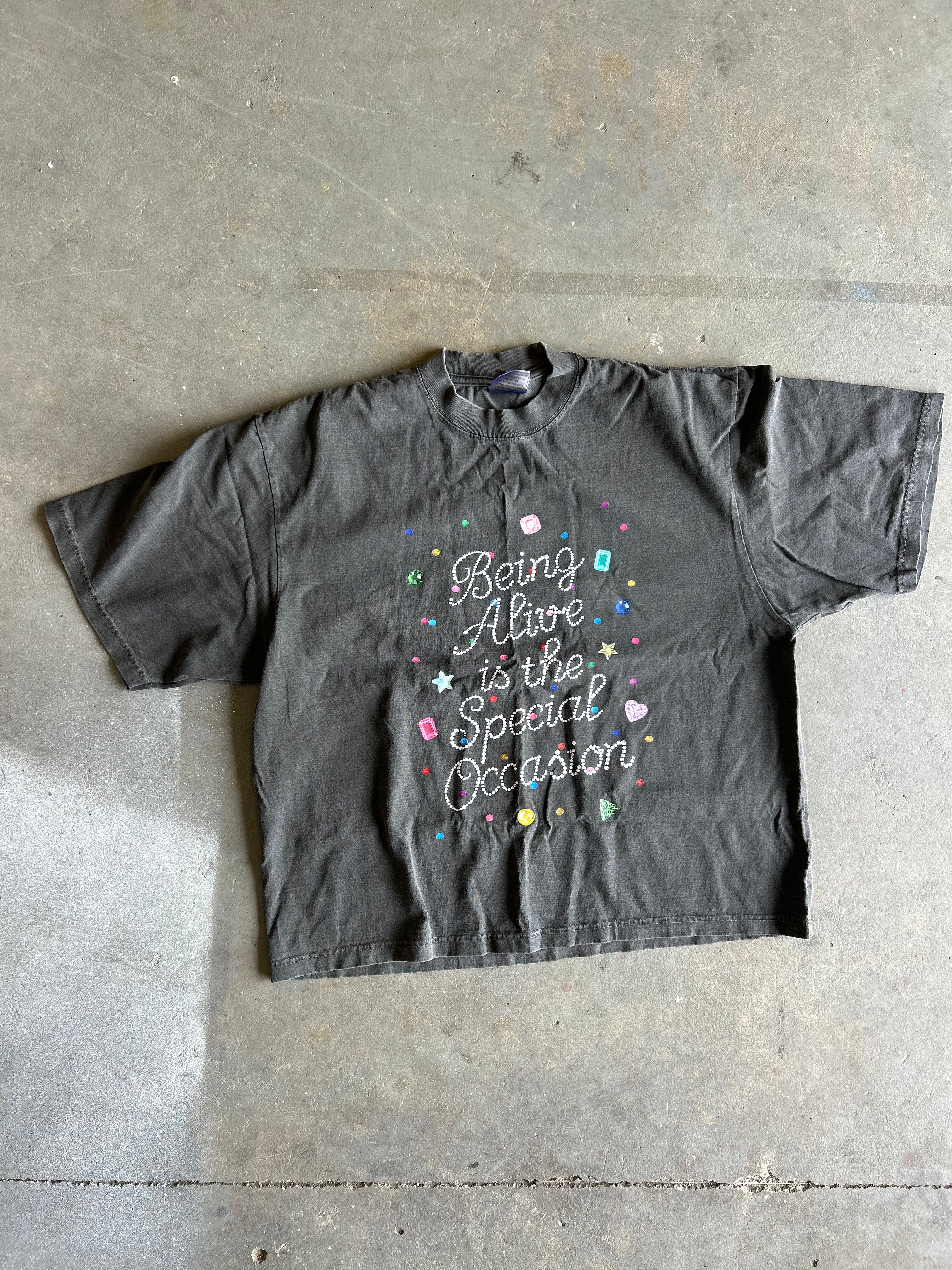 A faded black graphic t-shirt laid flat on a concrete floor, featuring the text 'Being Alive is the Special Occasion' in white script with colorful pastel glitter accents.