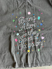 A close-up of a dark gray t-shirt featuring the phrase 'Being Alive is the Special Occasion' in white cursive rhinestone lettering, surrounded by colorful glittery star, heart, and dot embellishments.