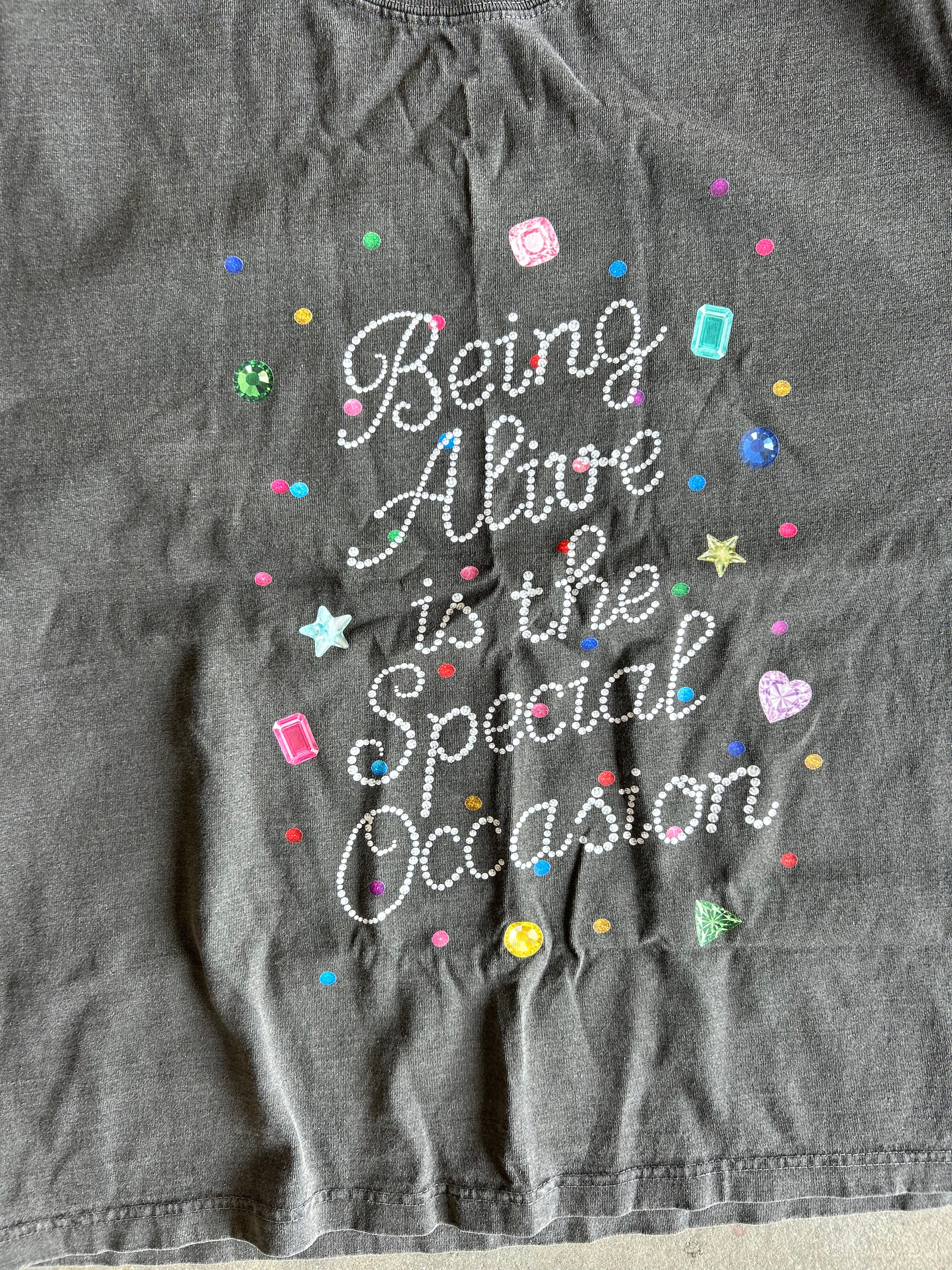 A close-up of a dark gray t-shirt featuring the phrase 'Being Alive is the Special Occasion' in white cursive rhinestone lettering, surrounded by colorful glittery star, heart, and dot embellishments.