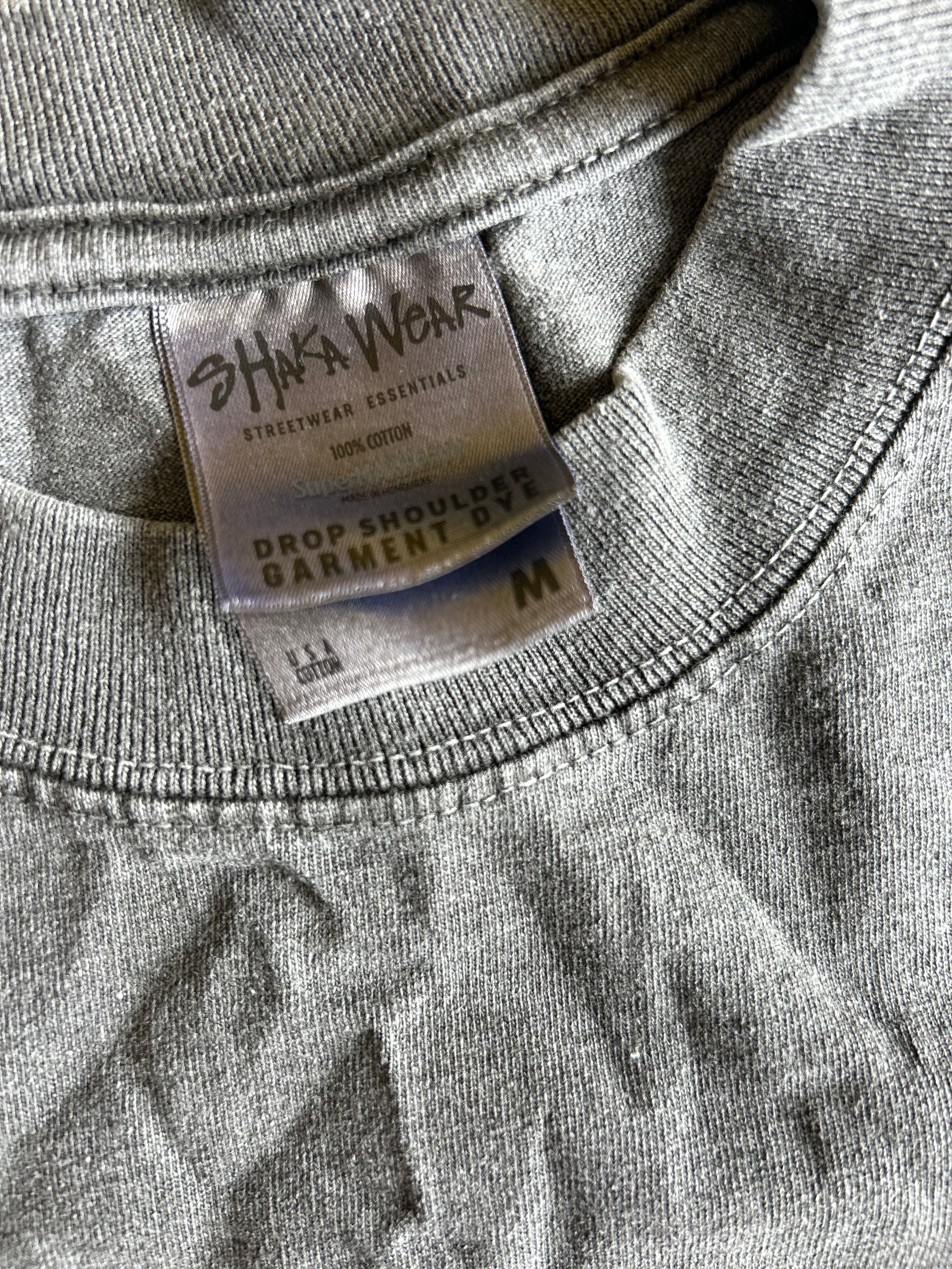 Close-up of a gray heather crewneck sweatshirt showing the inner neck label which reads 'SHAKA WEAR' and 'DROP SHOULDER GARMENT SWEATSHIRT' with size 'M'.