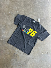 A faded black graphic t-shirt featuring a yellow '76' and a white script signature, laid flat on a textured concrete floor.