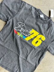 A dark gray washed t-shirt featuring a graphic print of the number 76 in yellow, alongside a white script logo reading 'Burnt Font' over a small red, green, and blue striped design, laid flat on a beige surface.