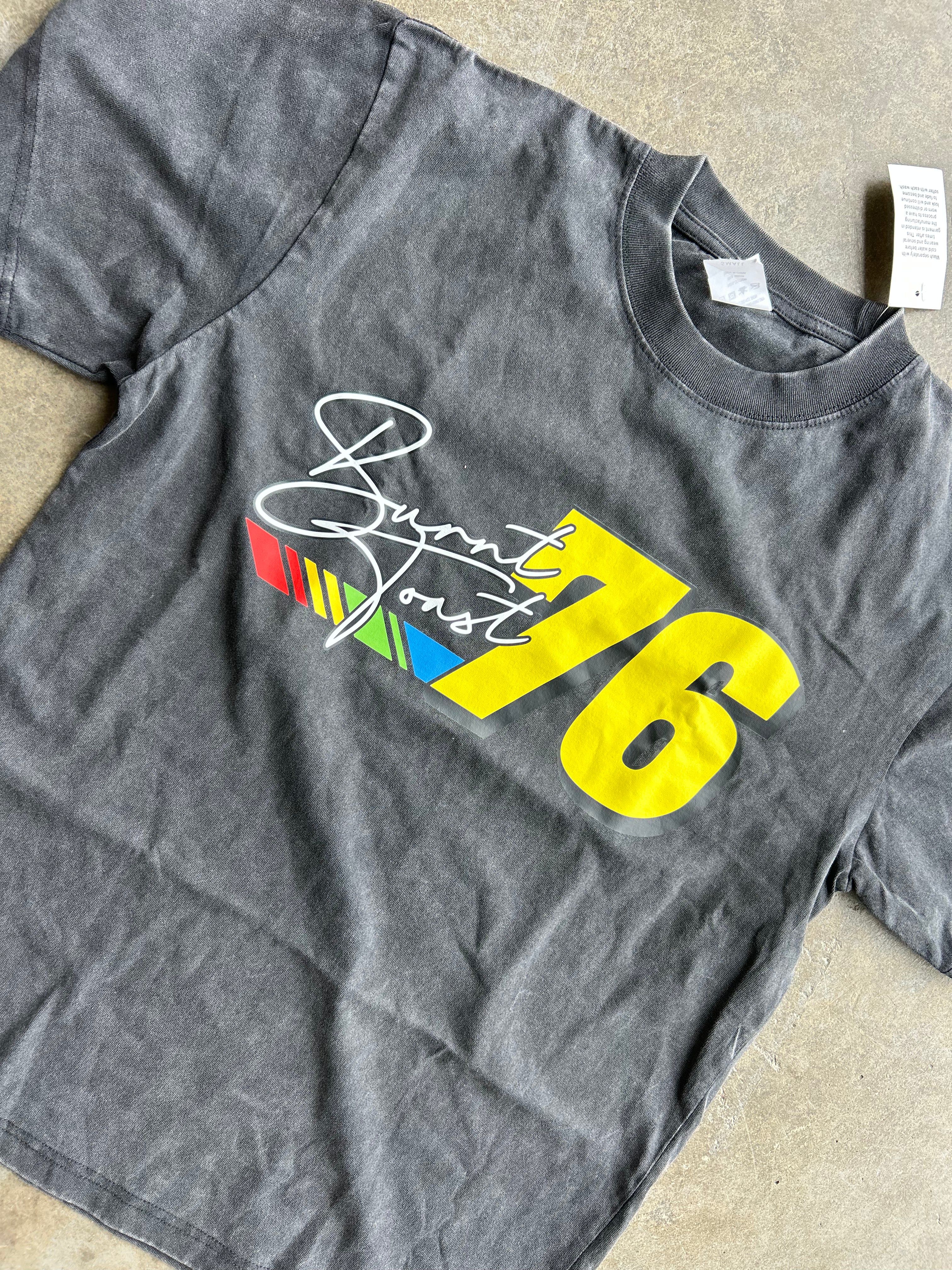 A dark gray washed t-shirt featuring a graphic print of the number 76 in yellow, alongside a white script logo reading 'Burnt Font' over a small red, green, and blue striped design, laid flat on a beige surface.