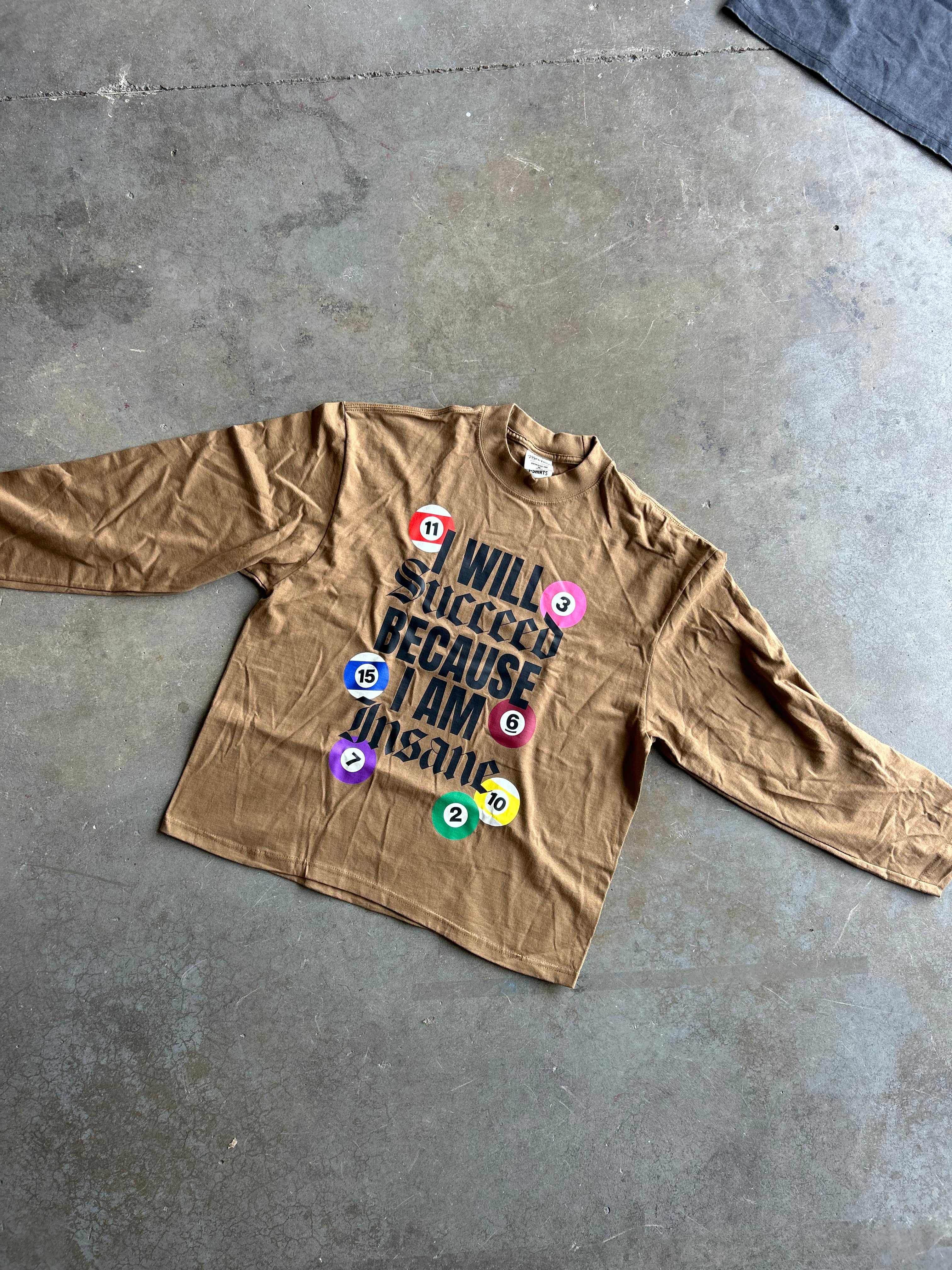 A tan long-sleeve graphic t-shirt laid flat on a gray concrete floor. The shirt features a bold black text graphic reading 'I WILL SUCCEED BECAUSE I AM INSPIRED' surrounded by colorful numbered balls (red 1, pink 2, orange 3, yellow 4, green 5, blue 6, purple 7).