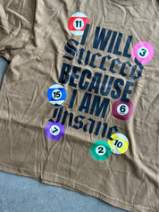 A close-up of a tan graphic t-shirt featuring the text 'I WILL Succeed BECAUSE I AM Insane' in dark blue gothic lettering, surrounded by colorful billiard ball graphics with numbers.