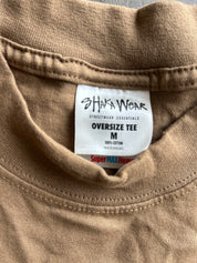Close-up of the inside collar of a tan oversized cotton t-shirt, showing a white brand label that reads 'SHAFAWEAR STREETWEAR ESSENTIALS OVERSIZE TEE M'.