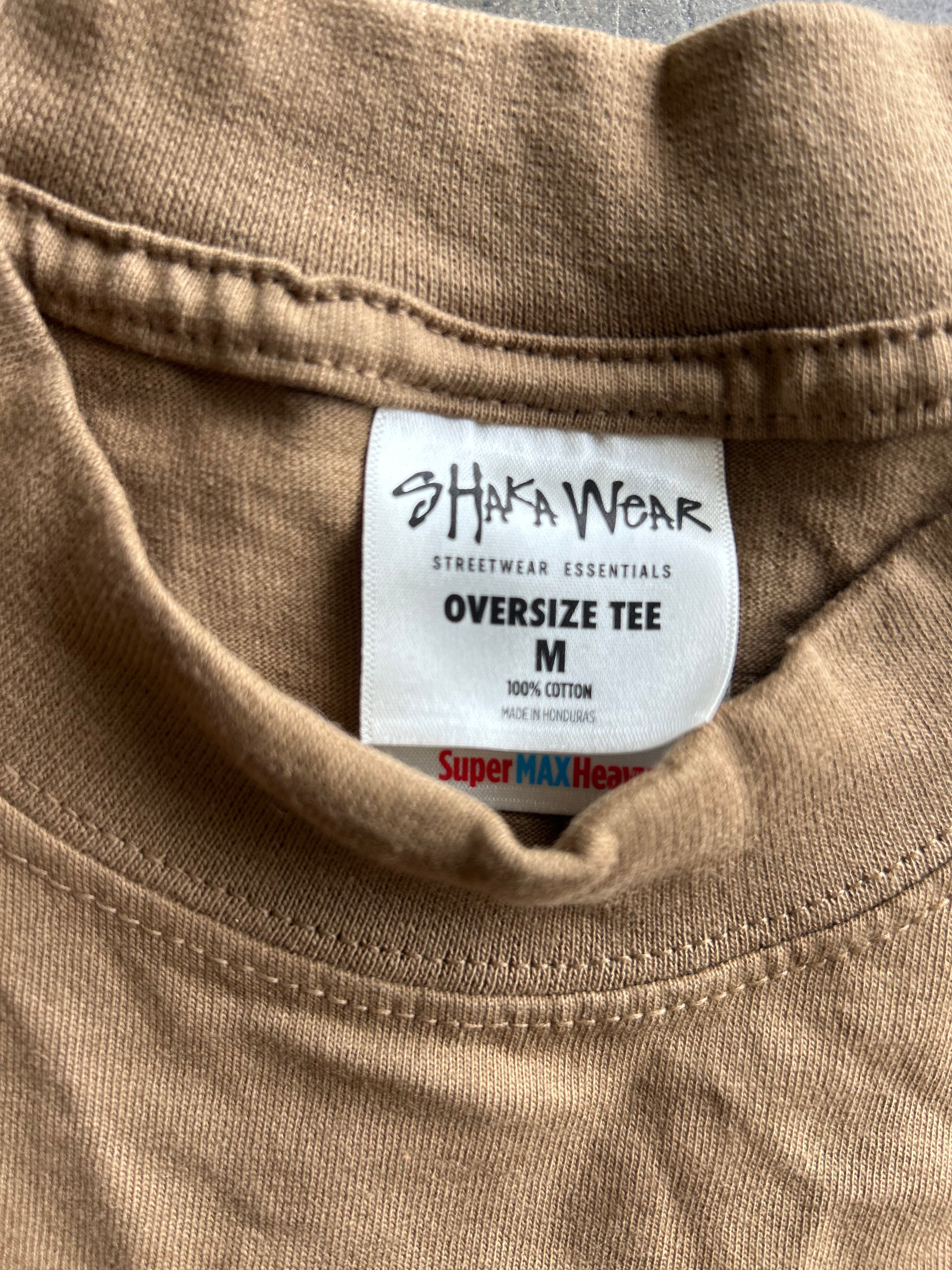 Close-up of the inside collar of a tan oversized cotton t-shirt, showing a white brand label that reads 'SHAFAWEAR STREETWEAR ESSENTIALS OVERSIZE TEE M'.