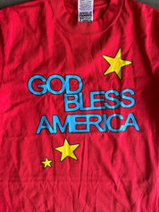 A red short-sleeve t-shirt featuring the text 'GOD BLESS AMERICA' in blue outline lettering with yellow stars, laid flat on a dark surface.