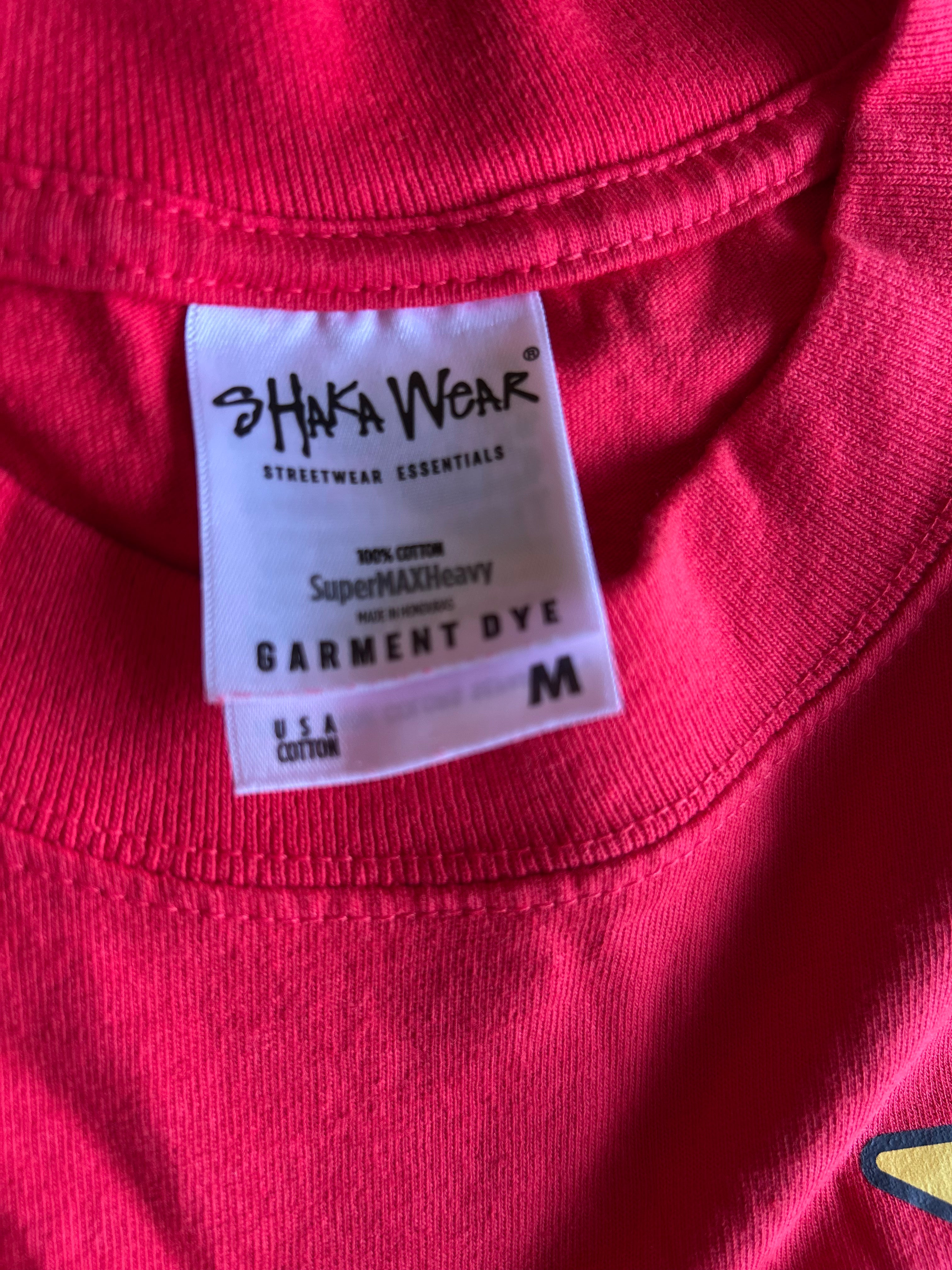 Close-up of a bright pink garment dye label reading 'SHAKA WEAR' and 'GARMENT DYE' on a red or pink t-shirt.