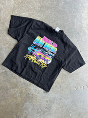 A black short-sleeve graphic t-shirt laid flat on a textured concrete floor. The shirt features a colorful retro-style print of a pink and blue muscle car with the text 'Cruisin' Man' and 'You Ain't Nothin' But' in neon pink, yellow, and blue lettering.