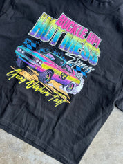 A black short-sleeve graphic t-shirt laid flat on a concrete floor. The shirt features a vibrant, multicolored graphic print of a classic race car with the text 'BUCKLE UP! HOT MESS' and 'Goody Owls Are Fast'.