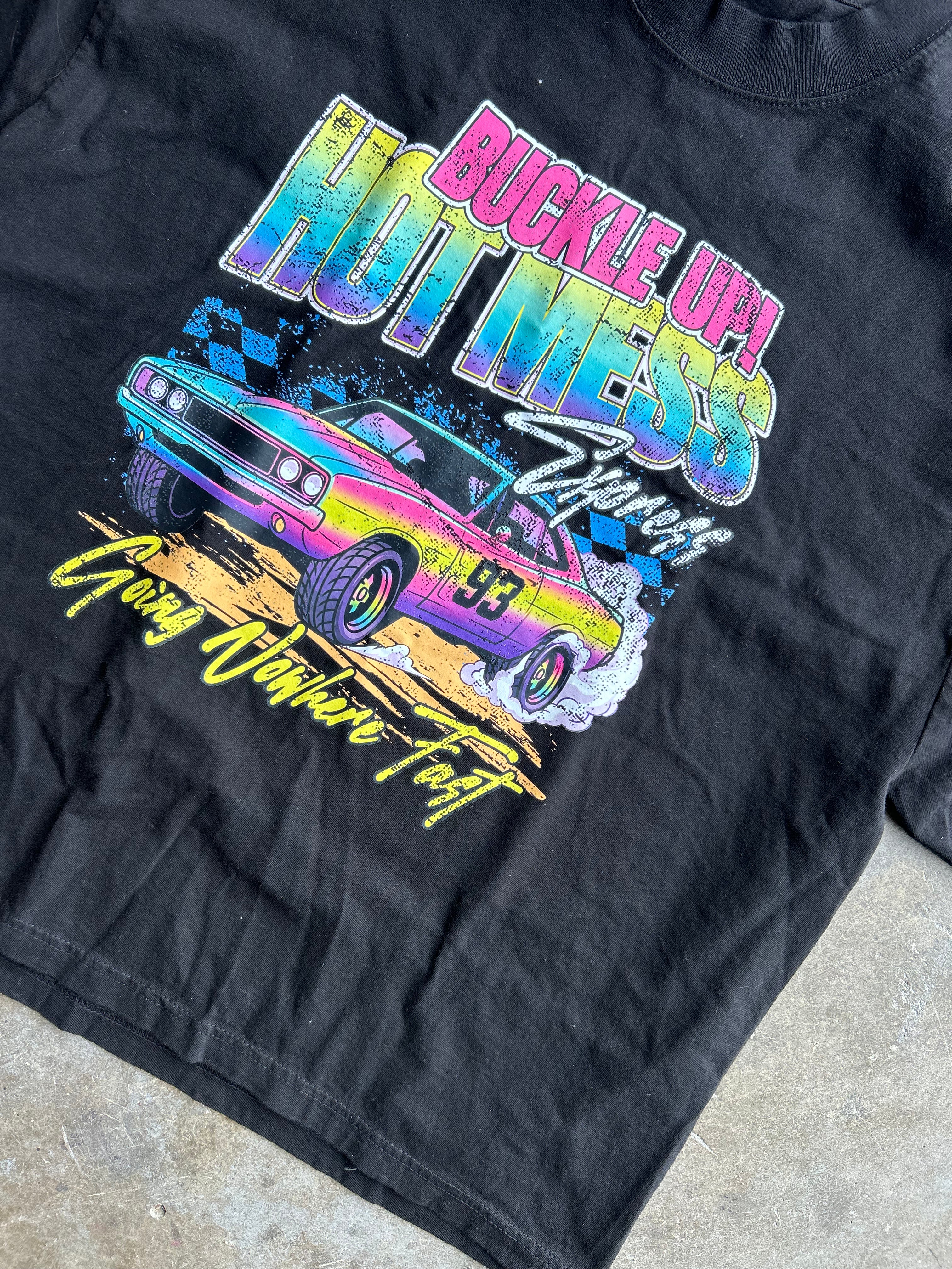A black short-sleeve graphic t-shirt laid flat on a concrete floor. The shirt features a vibrant, multicolored graphic print of a classic race car with the text 'BUCKLE UP! HOT MESS' and 'Goody Owls Are Fast'.