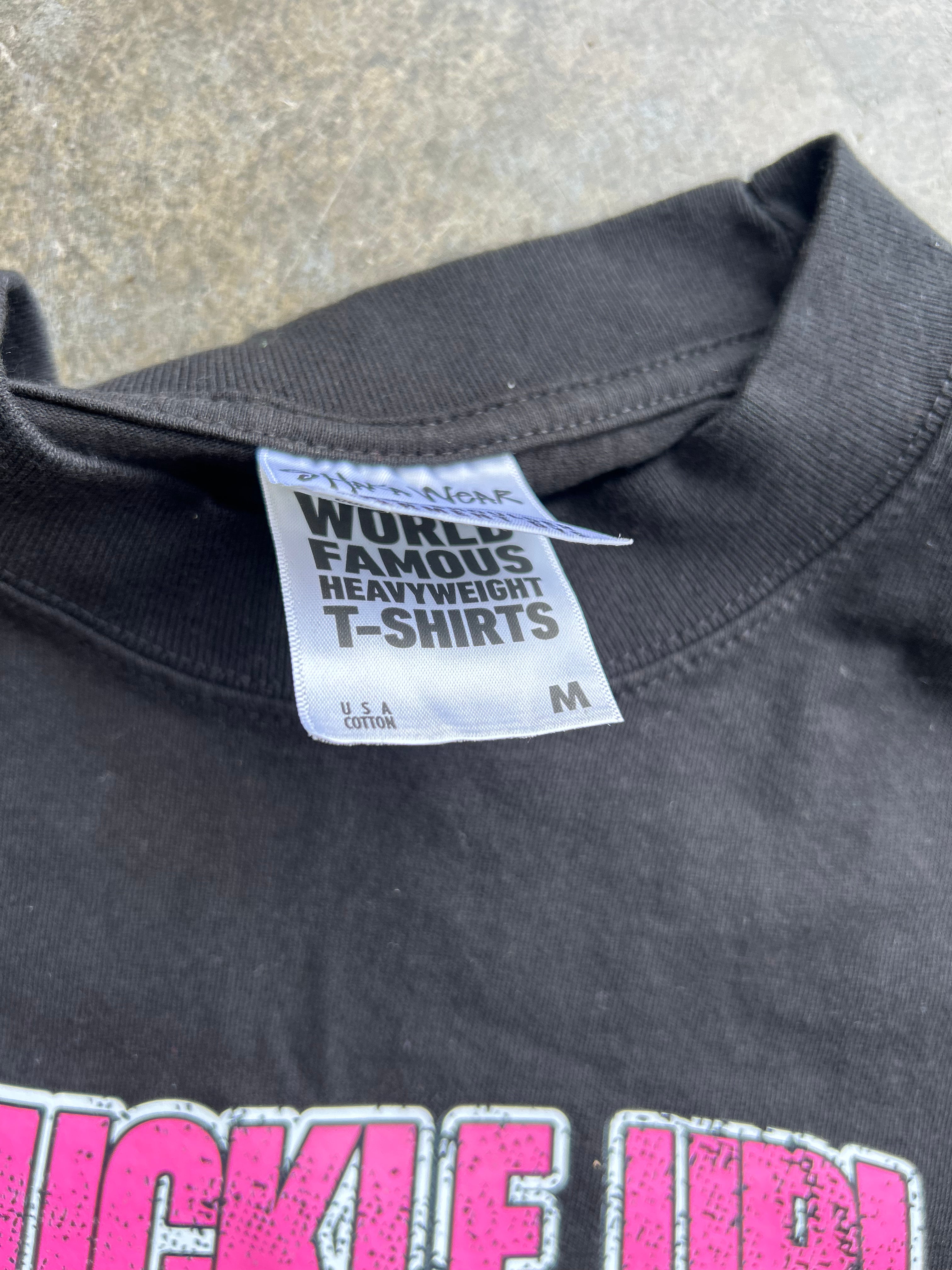 Close-up of the inside collar of a black heavyweight T-shirt, showing the white woven neck label that reads 'WORLD FAMOUS HEAVYWEIGHT T-Shirts' and 'M'. Part of a bright pink graphic print is visible at the bottom.