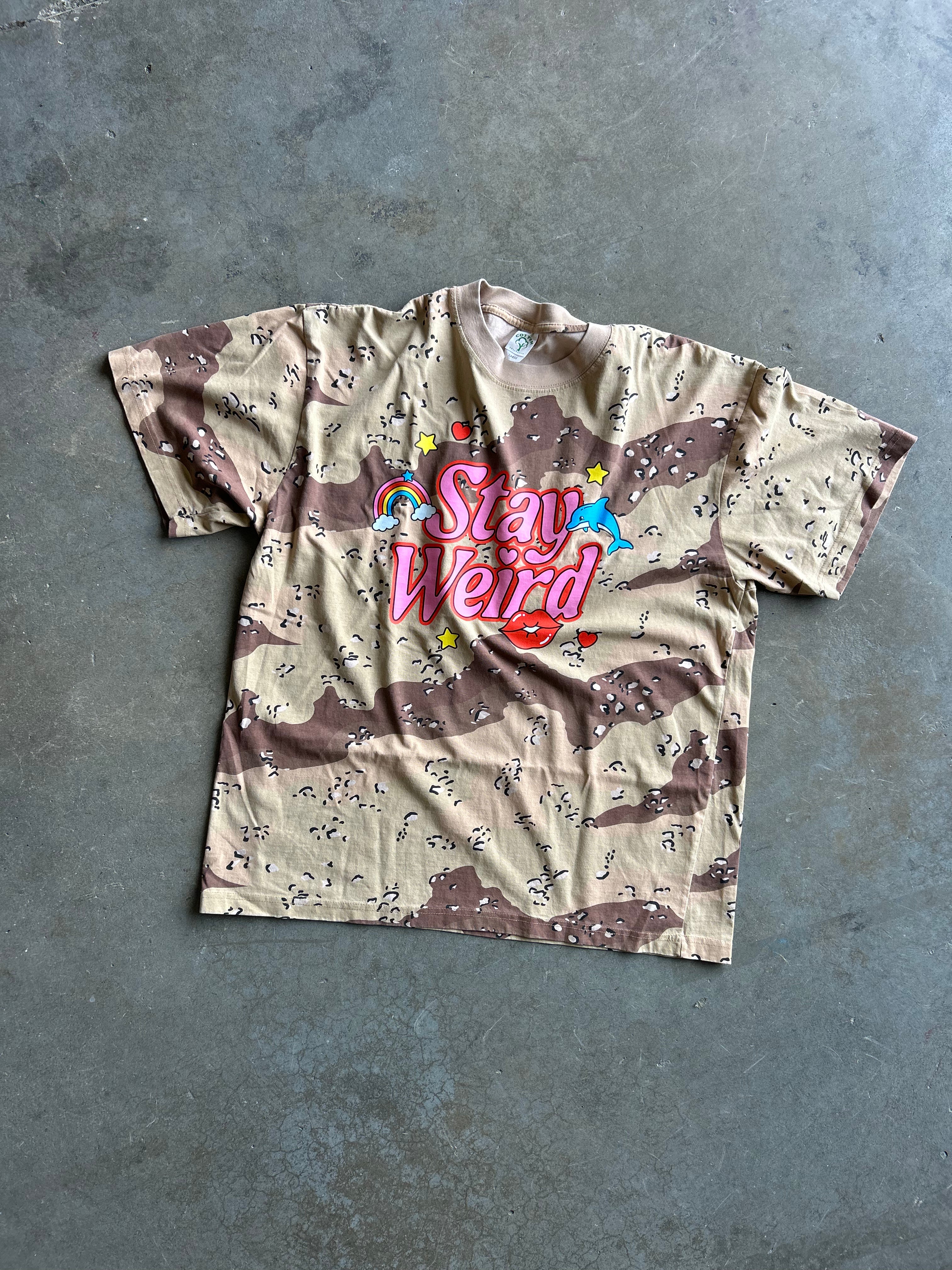 A short-sleeve graphic t-shirt with a beige and brown camouflage pattern, featuring a colorful 'Stay Weird' text graphic on the chest, laid flat on a gray concrete floor.