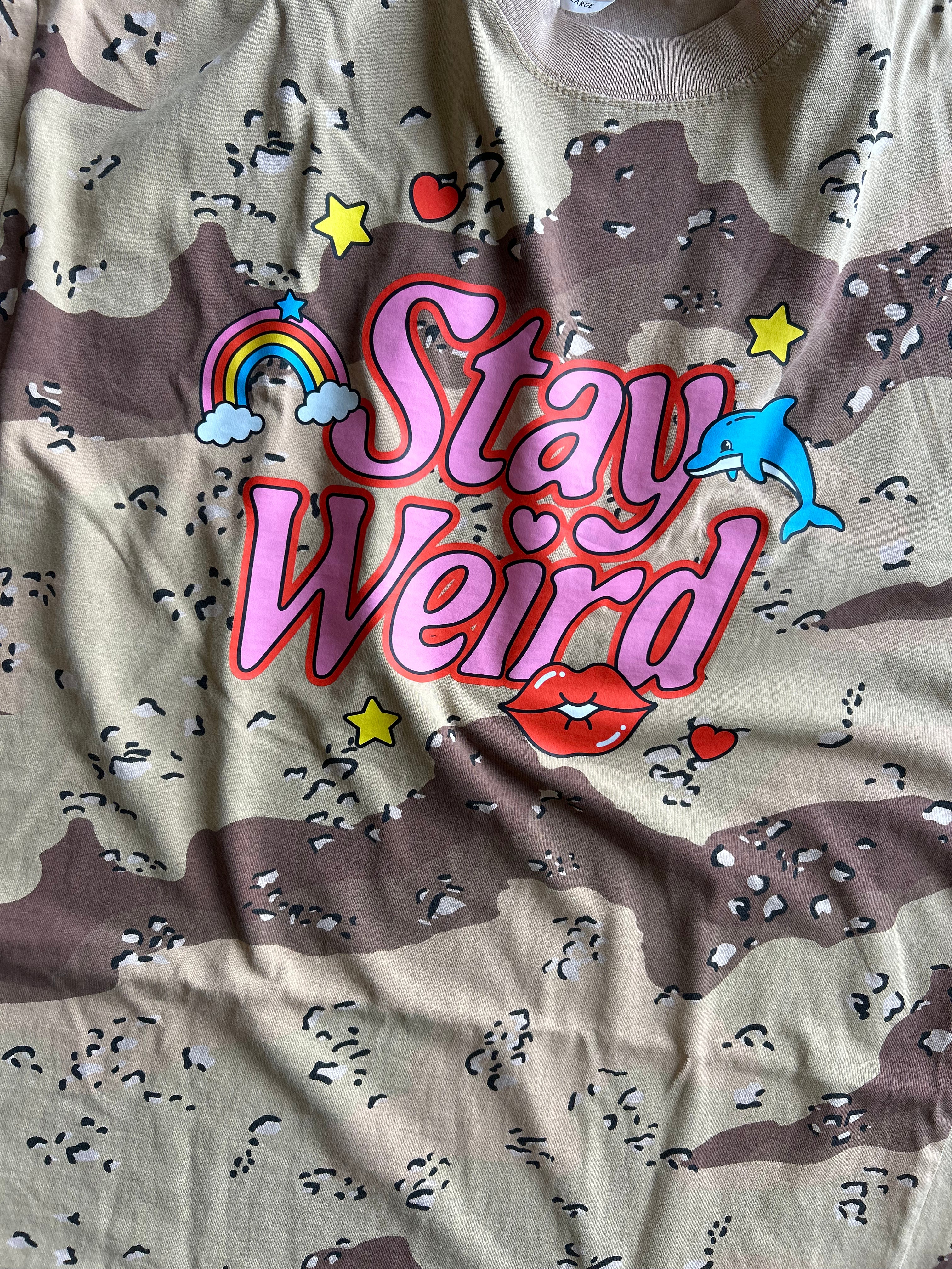 A close-up of a beige and brown camouflage t-shirt featuring a colorful graphic print that reads 'Stay Weird' in pink bubble letters, decorated with a rainbow, clouds, stars, a blue dolphin, red lips, and small hearts.