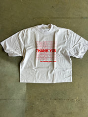 A white oversized short-sleeve t-shirt with a red graphic text print reading 'THANK YOU for raising your hand' in a stacked, layered design, laid flat on a concrete floor.