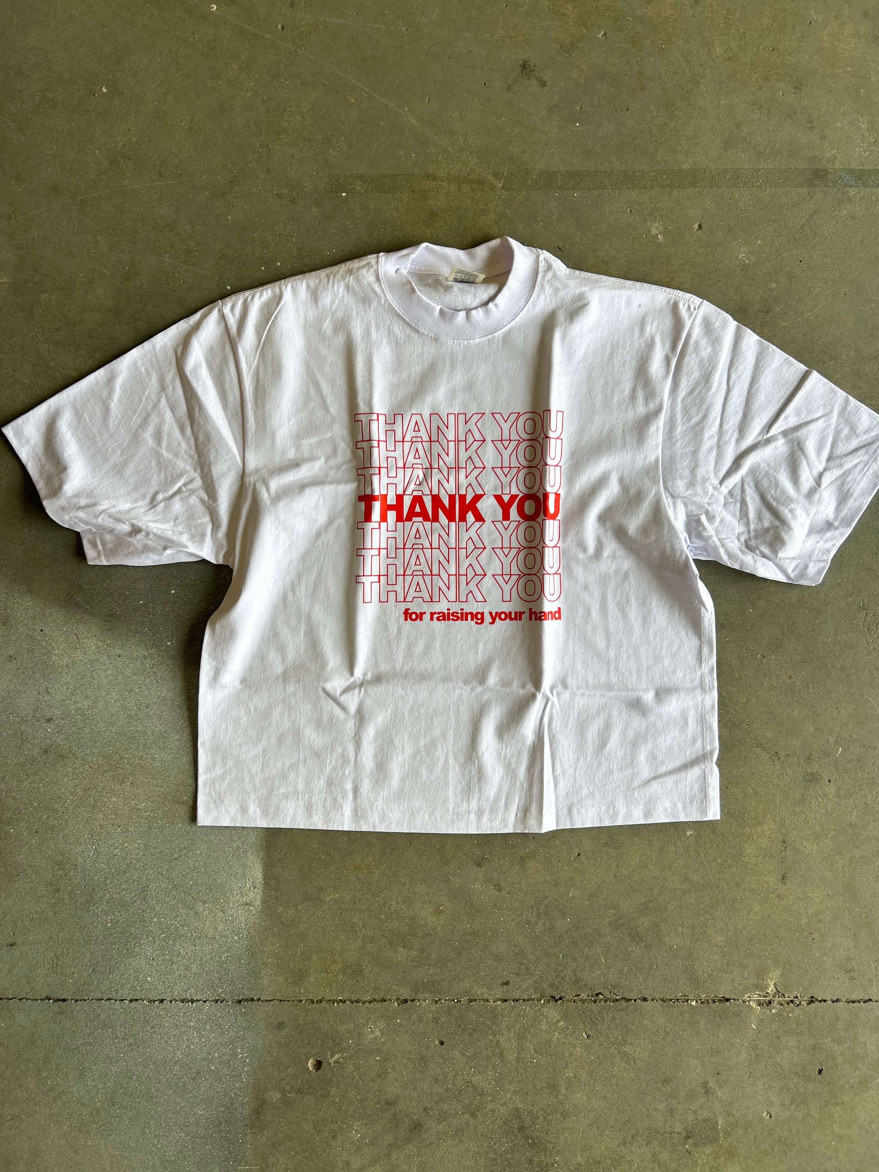 A white oversized short-sleeve t-shirt with a red graphic text print reading 'THANK YOU for raising your hand' in a stacked, layered design, laid flat on a concrete floor.