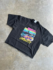 A black short-sleeve graphic t-shirt laid flat on a textured concrete floor. The shirt features a colorful retro-style print of a race car with neon pink, yellow, and blue lettering that reads 'Racing Hotness' and 'Fast Lane Forever'.