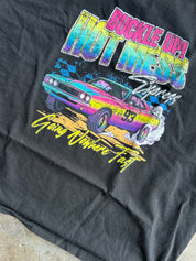 Close up of a black short-sleeve graphic t-shirt laid flat on a textured concrete floor. The shirt features a colorful retro-style print of a race car with neon pink, yellow, and blue lettering that reads 'Racing Hotness' and 'Fast Lane Forever'.