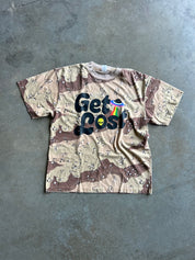 A short-sleeve crewneck t-shirt with a beige and brown desert camouflage pattern, featuring a graphic on the front that reads 'Get Lost' in black bubble letters with a small rainbow flag and alien icon.