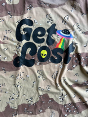 A close-up of a camouflage t-shirt featuring a graphic print that reads 'Get Lost' in black bubble letters. The letter 'o' is replaced by a yellow alien head, and the 'st' is replaced by a colorful rainbow-striped UFO.