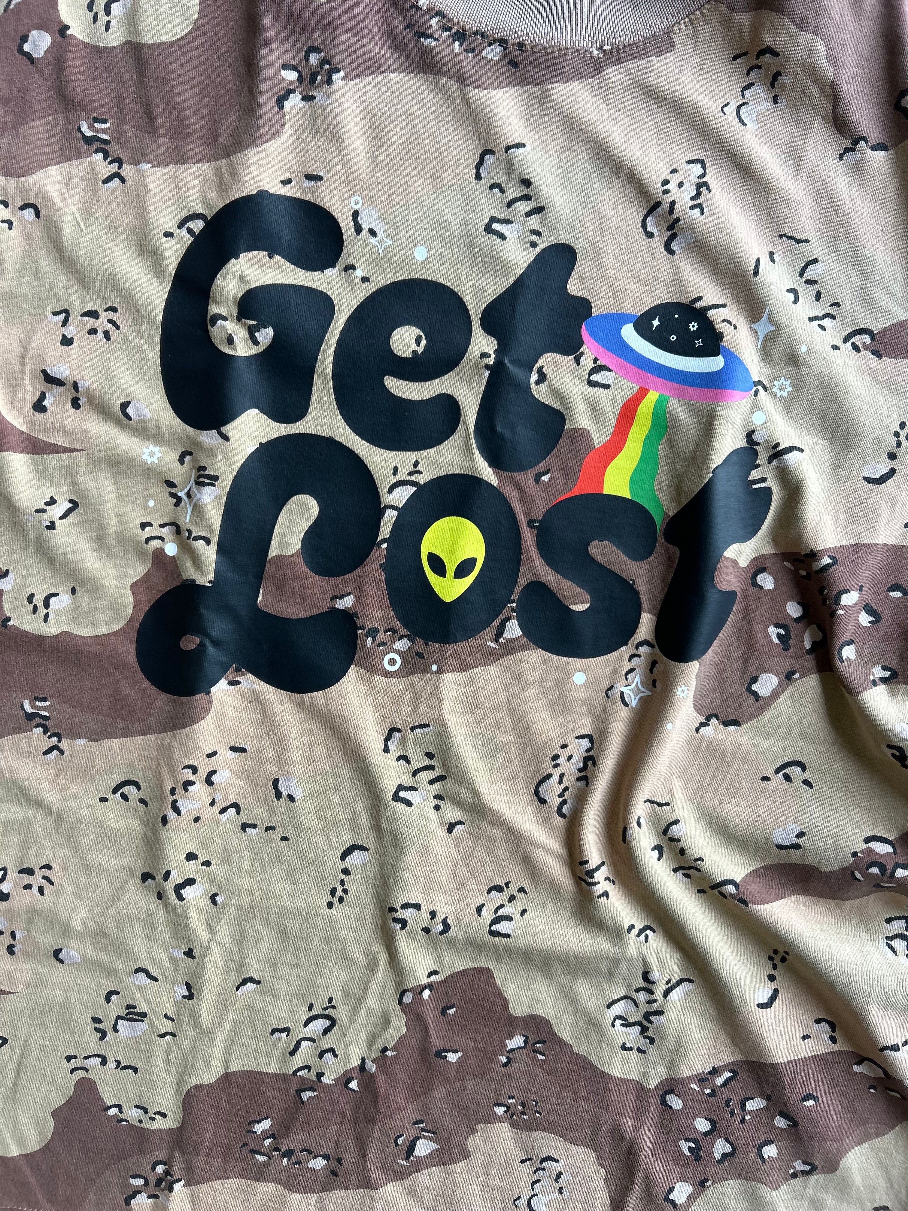 A close-up of a camouflage t-shirt featuring a graphic print that reads 'Get Lost' in black bubble letters. The letter 'o' is replaced by a yellow alien head, and the 'st' is replaced by a colorful rainbow-striped UFO.